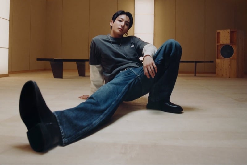 Jung Kook sits on the floor in blue jeans and black boots, pairing a gray tee with a white long-sleeve top.
