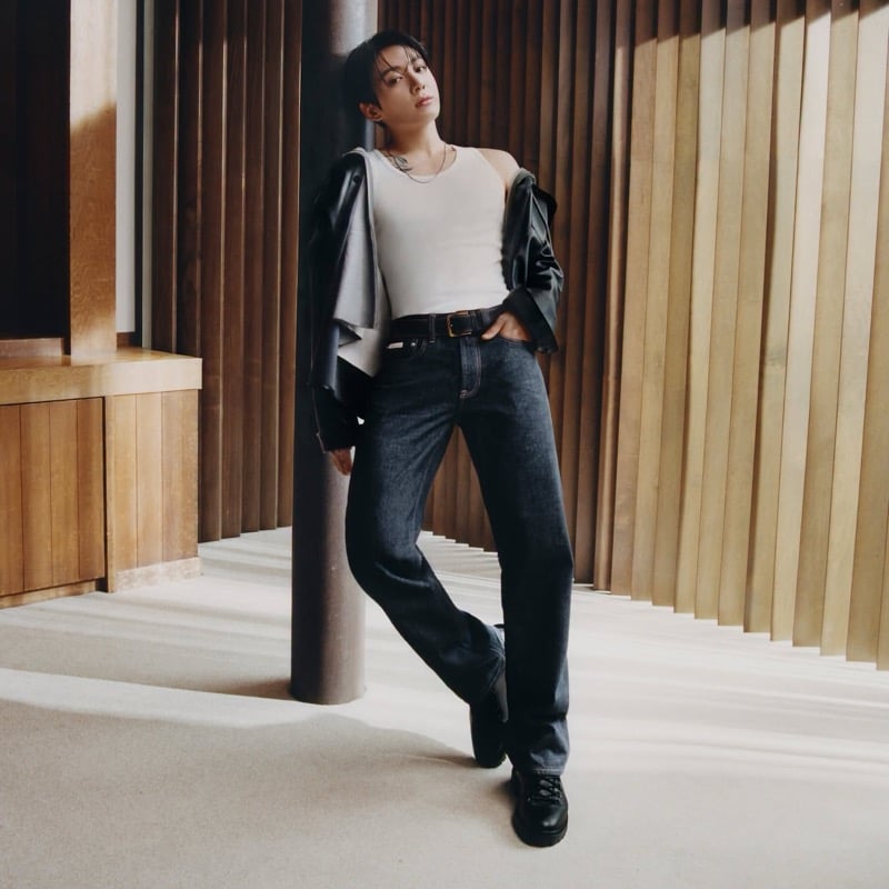 Jung Kook leans against a column in dark jeans, a white tank, and a black leather jacket.