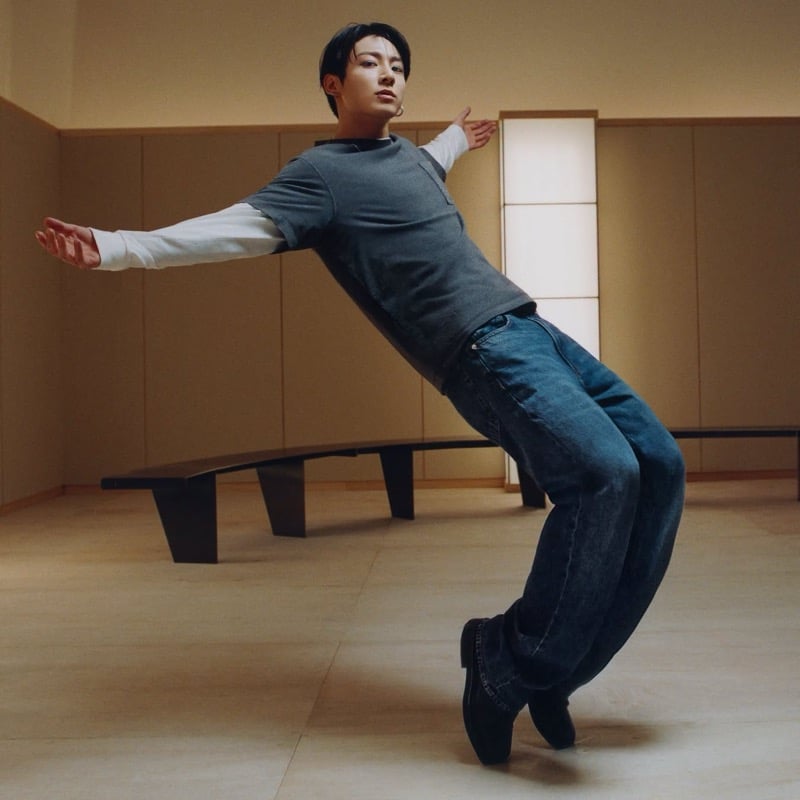 Jung Kook leans back with his arms outstretched in layered tees, blue jeans, and black boots.