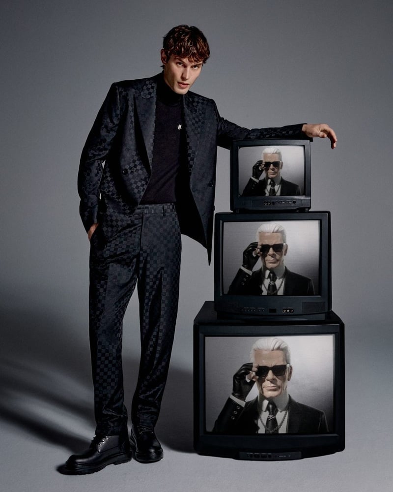 Kit Butler wears a dark checkerboard suit and black turtleneck beside stacked televisions showing NOT-KARL.