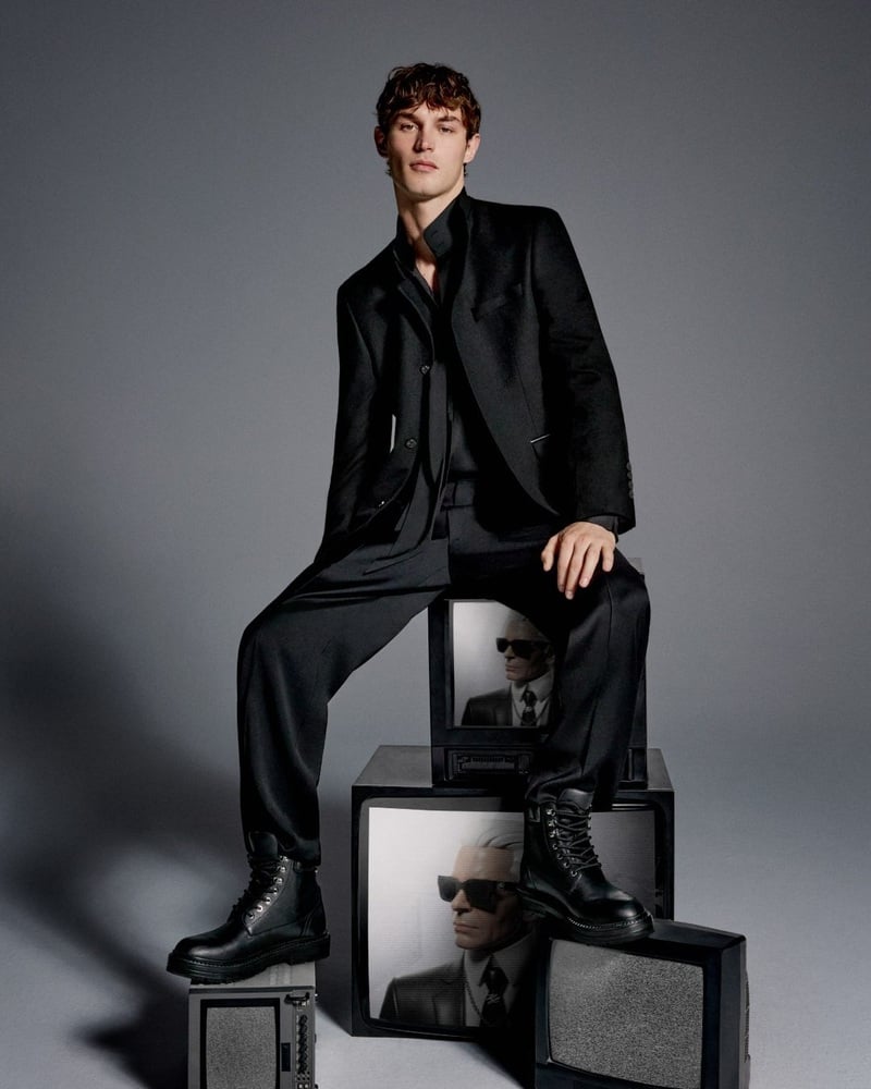 Kit Butler sits on stacked televisions wearing a black suit, open-neck shirt, and lace-up boots.