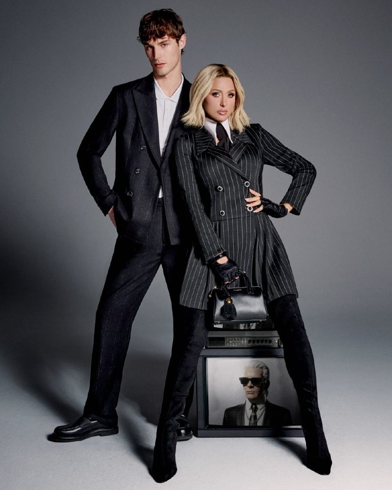Kit Butler stands beside Paris Hilton in dark tailoring for Karl Lagerfeld, with Hilton wearing a pinstriped suit.