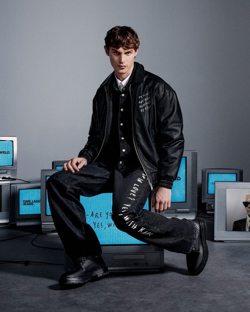 Kit Butler wears a black jacket and dark trousers with white lettering beside a group of televisions.