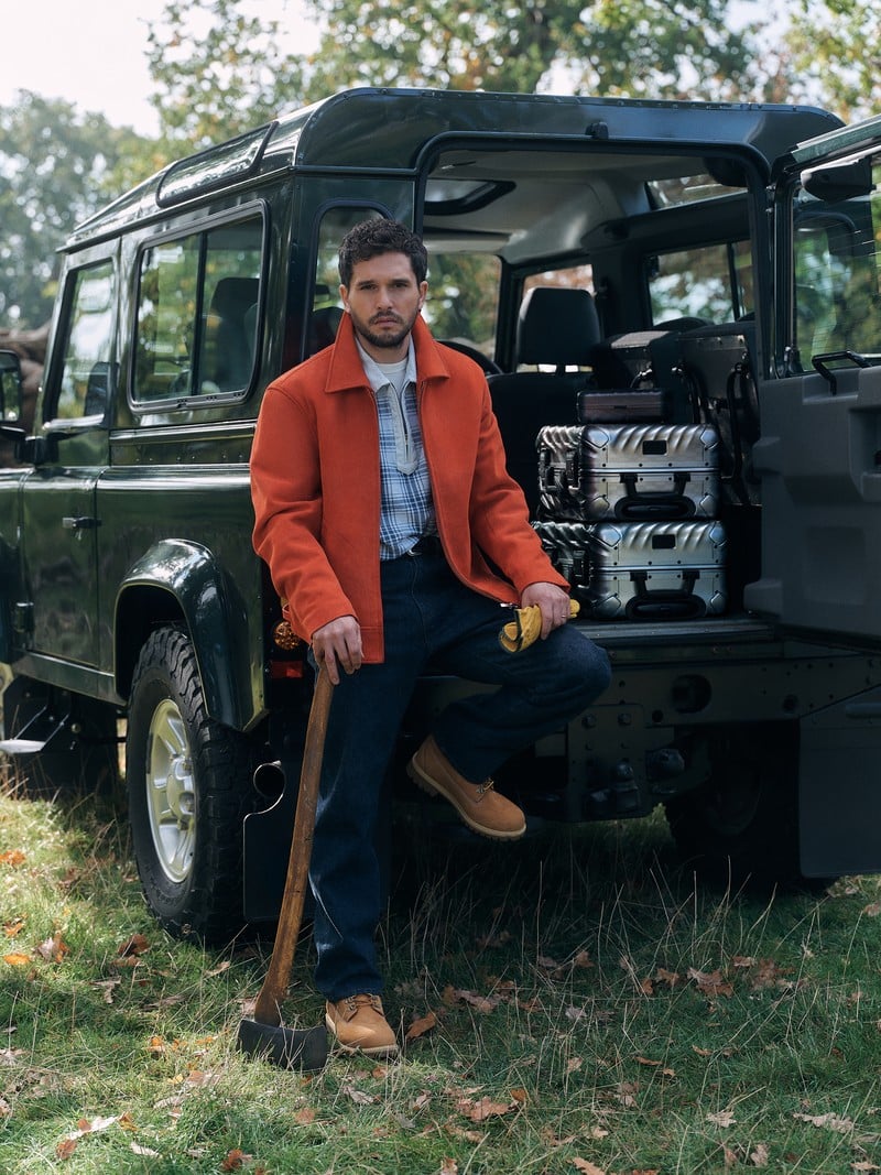 Kit Harington wears an orange jacket and jeans beside an off-road vehicle.