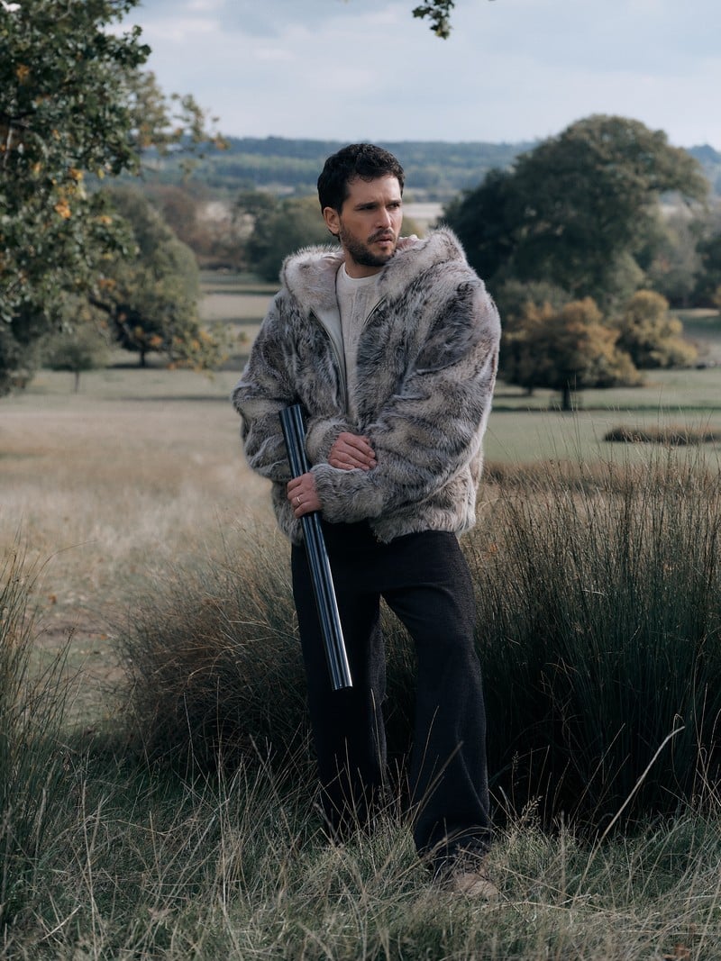 Kit Harington wears a hooded gray shearling jacket while holding a shotgun in the countryside.