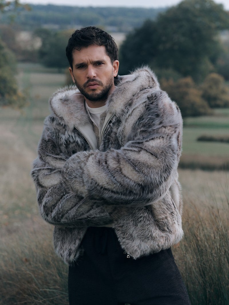 Kit Harington wears a hooded shearling jacket over dark trousers in a field.