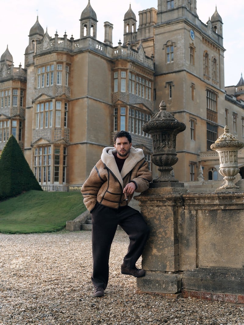 Kit Harington wears a tan shearling jacket and dark trousers outside a country house.