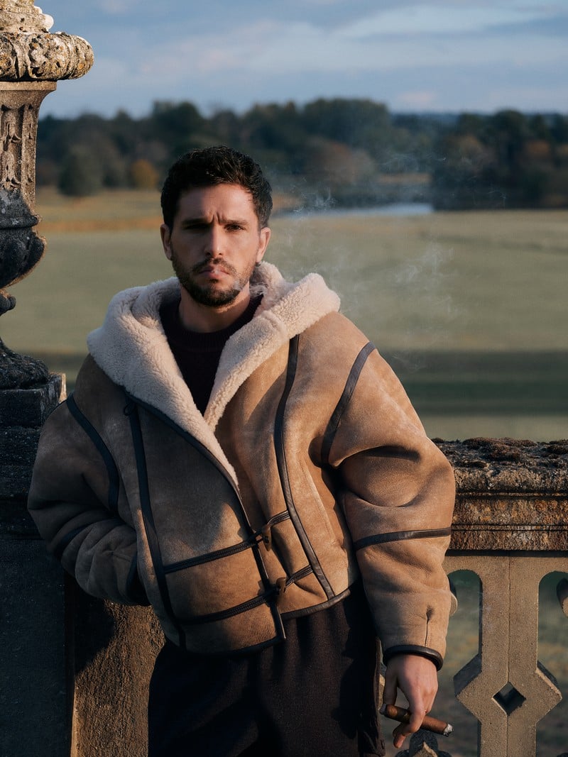 Kit Harington wears a tan shearling jacket with dark leather trim on a stone terrace.