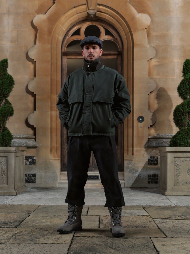 Kit Harington wears a dark jacket, flat cap, and lace-up boots outside a country house.
