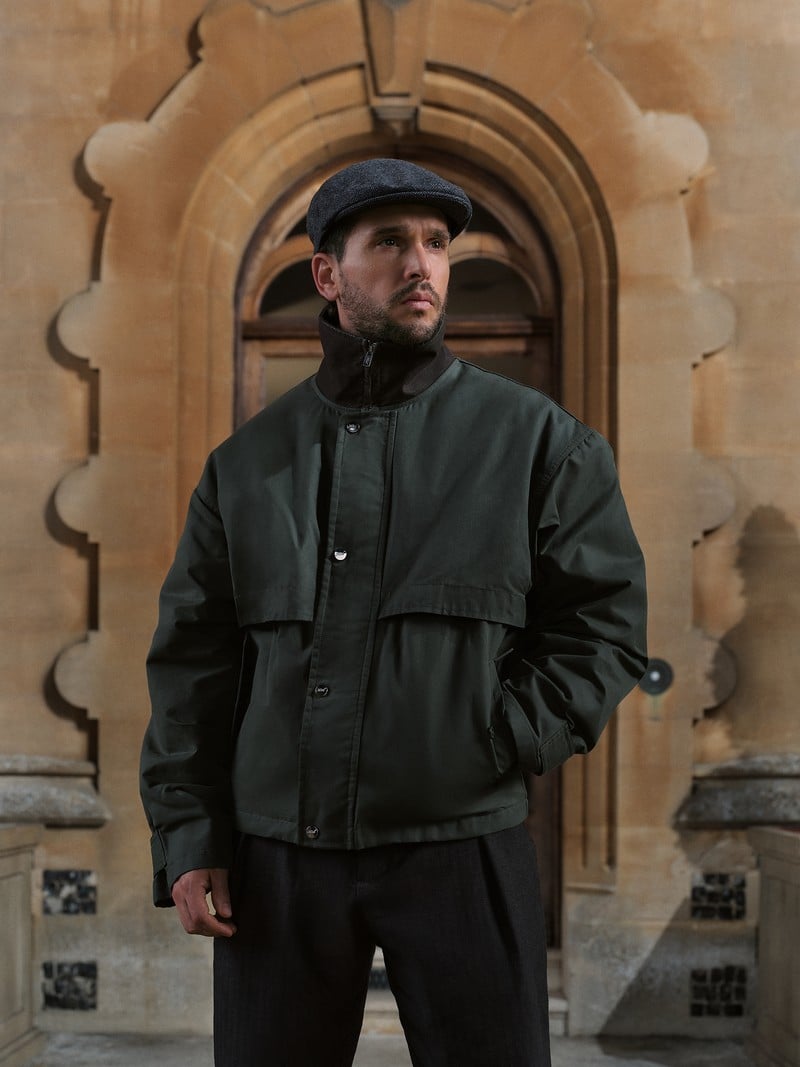 Kit Harington wears a dark high-collared jacket and flat cap outside a stone doorway.