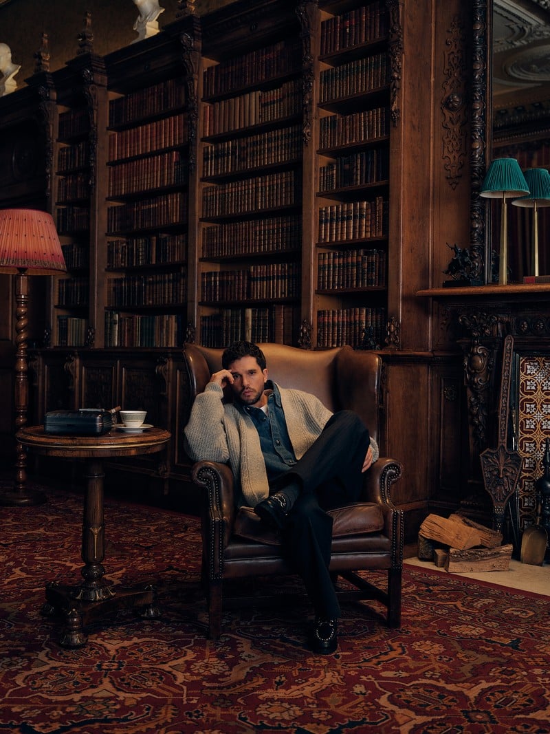 Kit Harington relaxes in a leather armchair wearing a cardigan and dark trousers in a library.