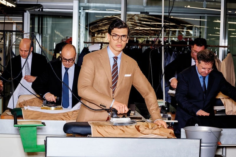 Atila Escolano Sanchez holds an iron at a workshop pressing table in a tan Kiton suit and striped tie.