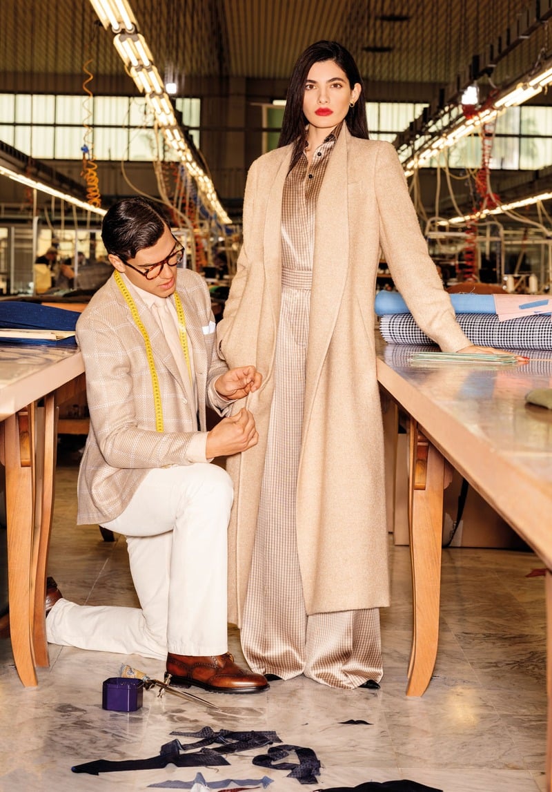 Atila Escolano Sanchez kneels beside Sara Fasano, who wears a long pale coat over a flowing outfit, in a tailoring workshop.