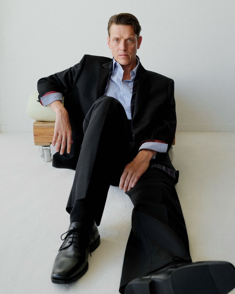 Sebastian Warschow sits in a black suit and blue striped shirt with one leg extended toward the camera.