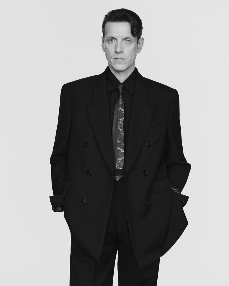 Black-and-white portrait of Sebastian Warschow in an oversized black double-breasted suit and paisley tie, with his hands in his pockets.