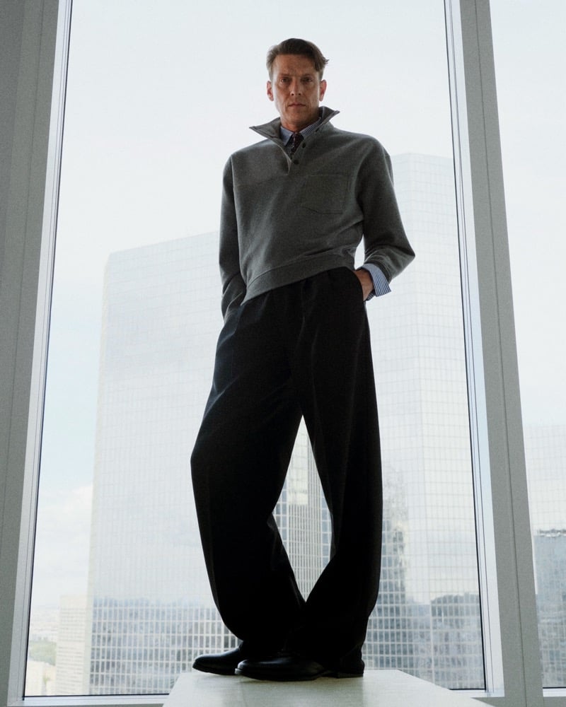 Sebastian Warschow stands by a tall window in a gray pullover over a blue striped shirt, wide-leg black trousers, and black shoes.