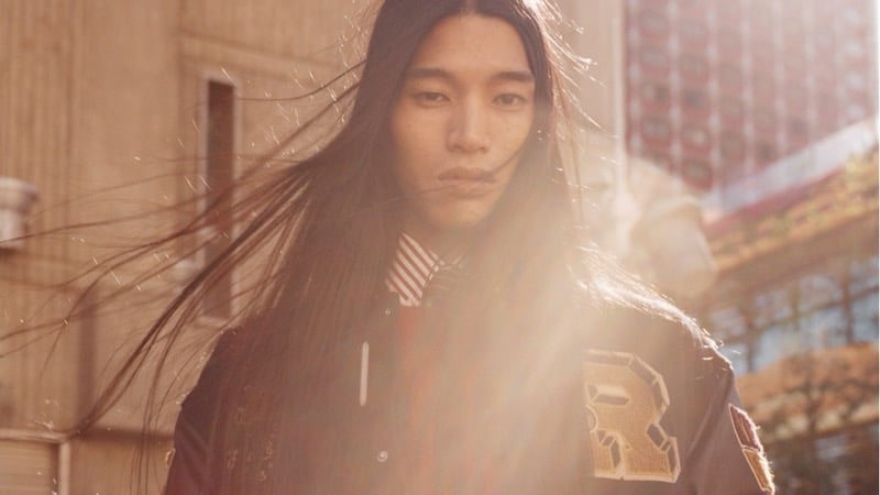 Kyosuke Akiyoshi with long hair blowing across his face in warm sunlight, wearing a varsity jacket with a gold letter patch over a striped shirt and tie