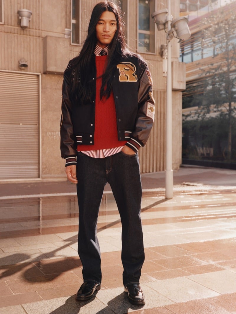 Kyosuke Akiyoshi standing on a sunlit plaza in a varsity jacket with leather sleeves and a gold letter patch over a red cable-knit sweater, striped shirt, tie, dark jeans, and tassel loafers