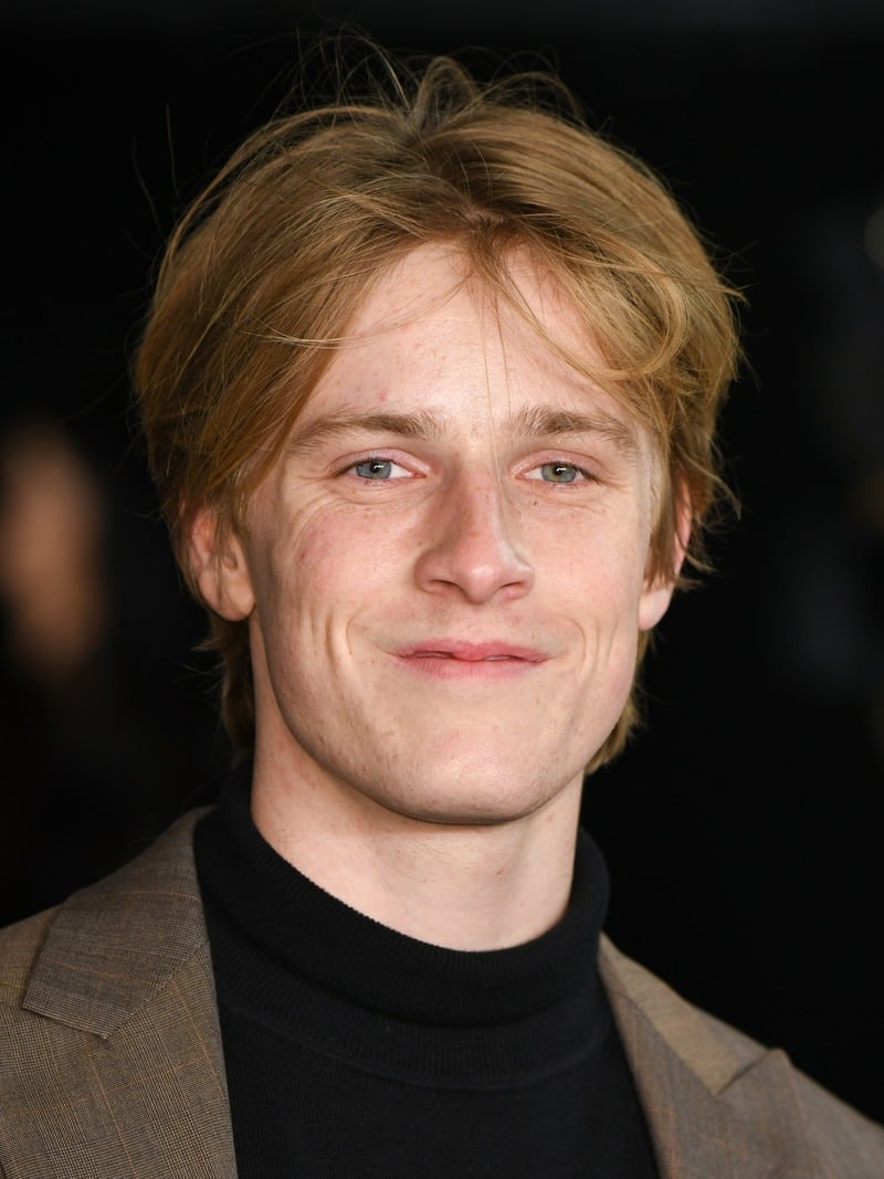 Louis Hofmann with loosely parted dark blonde hair falling across his forehead, wearing a brown jacket over a black turtleneck.
