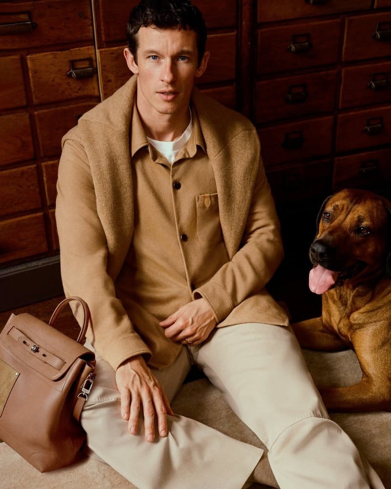 Callum Turner sits beside a dog with a Louis Vuitton bag, wearing a tailored jacket and matching shirt for the Trunk Edition campaign.