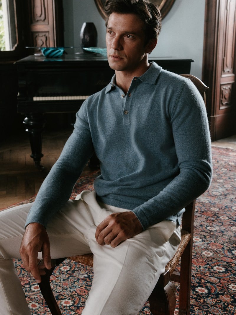 A man sits wearing a blue-gray quarter-zip sweater and white trousers.