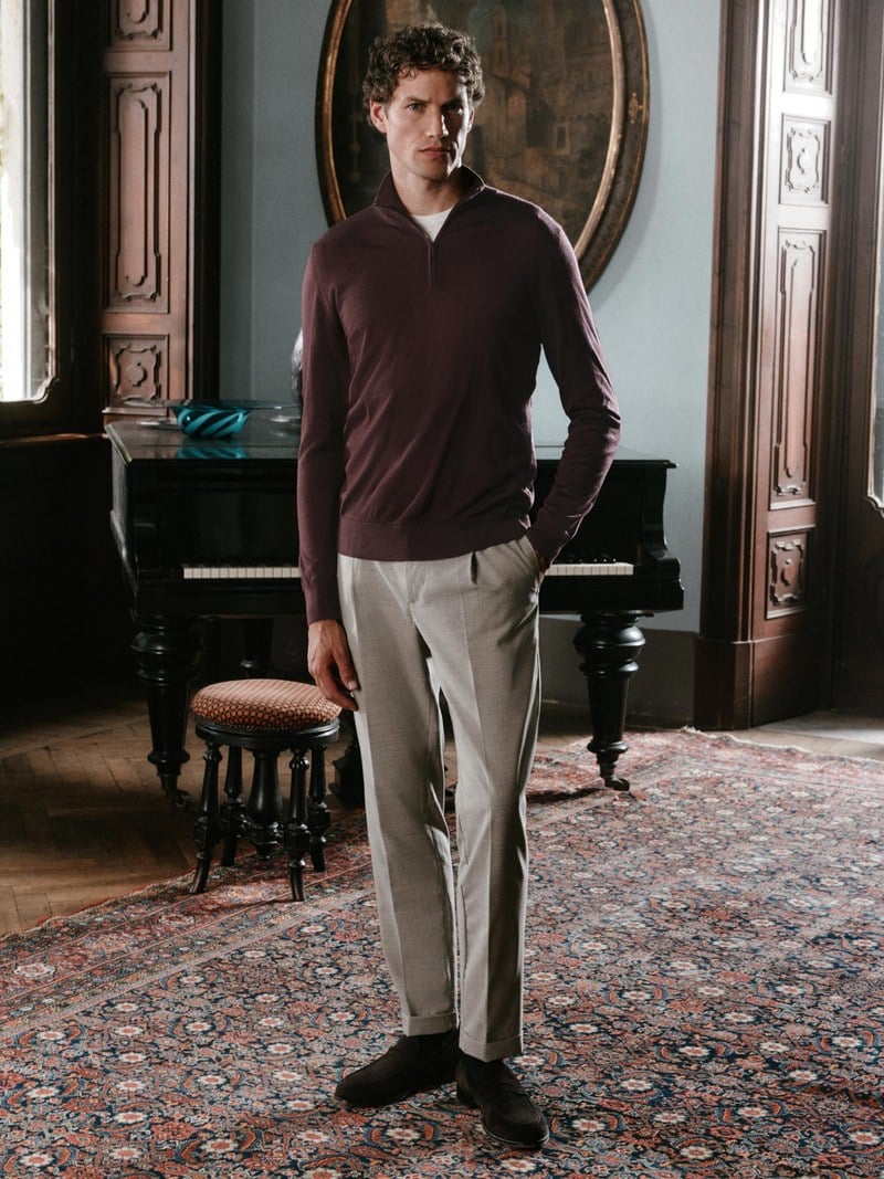A man stands in a burgundy V-neck sweater over a white shirt with light trousers.