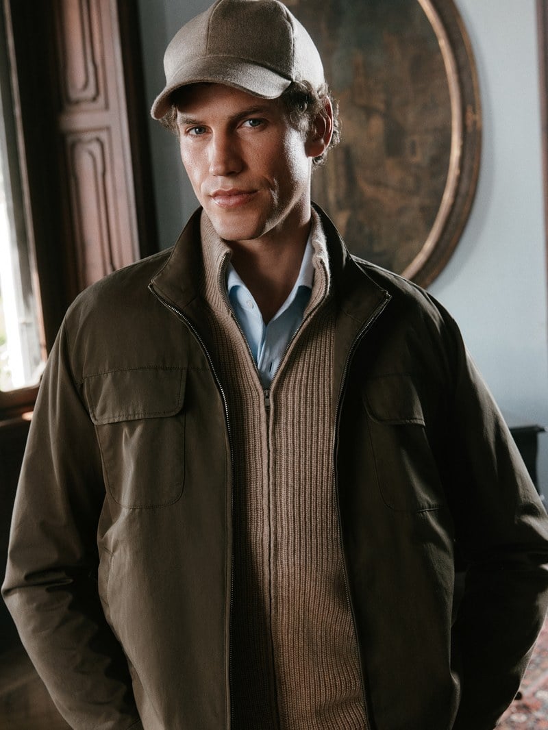 A man wears a muted olive jacket over a pale blue shirt and a matching cap.