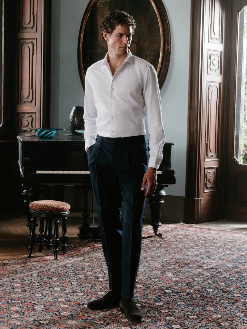 A man wears a white button-down shirt with dark tailored trousers.