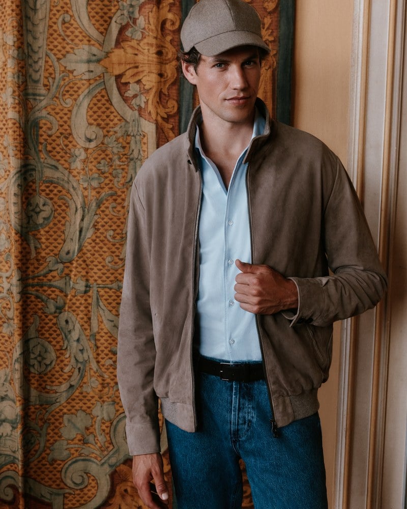 A man wears a taupe zip jacket over a pale blue shirt and a gray cap.