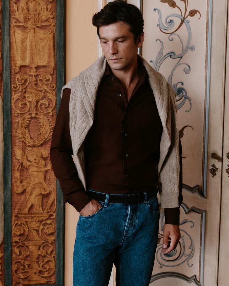 A man wears a dark brown shirt with a light sweater draped over his shoulders and blue jeans.