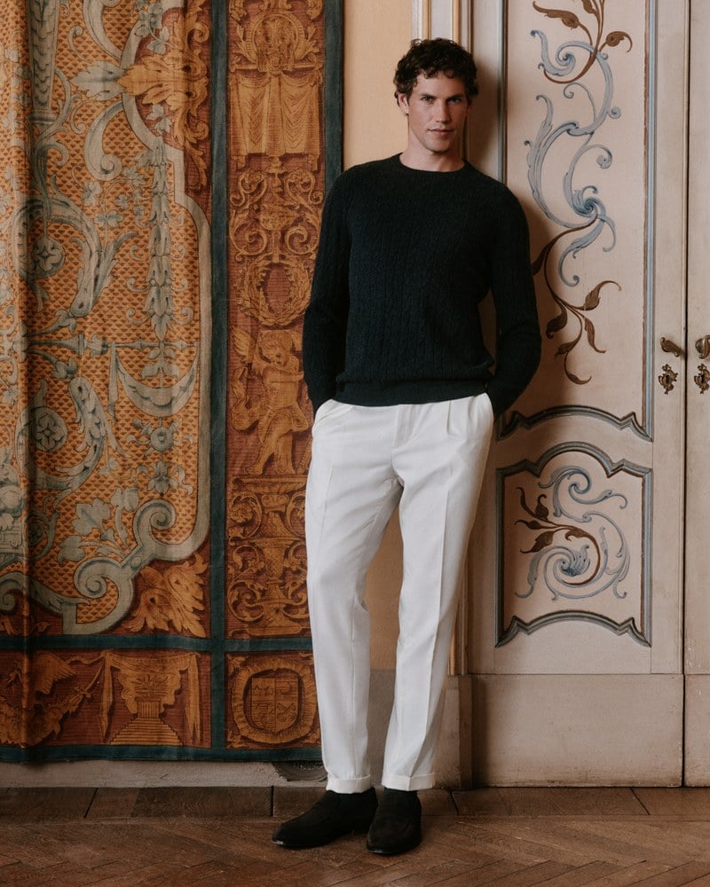 A man wears a black crewneck sweater with white trousers.