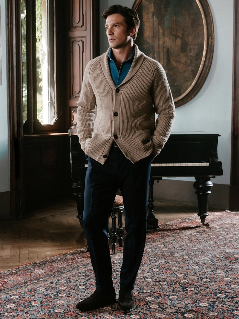 A man wears a taupe zip cardigan over a blue shirt and tie with dark trousers.
