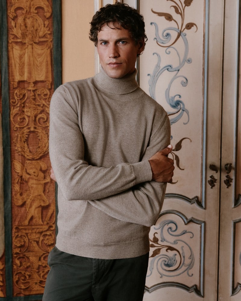 A man wears a beige crewneck sweater with dark trousers.
