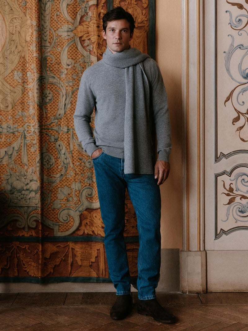 A man wears a light gray-blue crewneck sweater over a pale blue shirt with blue jeans.