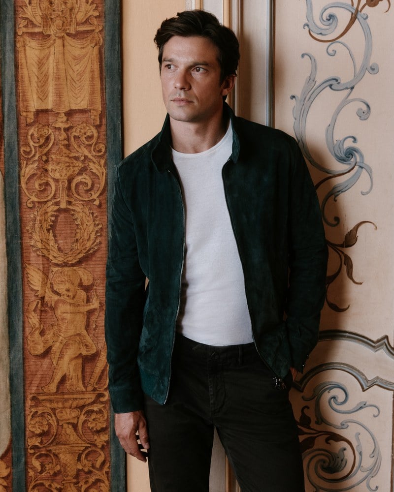 A man wears a dark green jacket over a white T-shirt with black trousers.