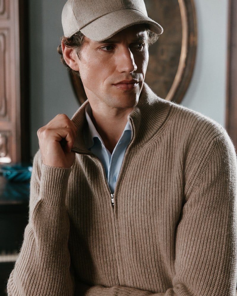 Close portrait of a man wearing a beige zip cardigan over a pale blue shirt and a light gray cap.