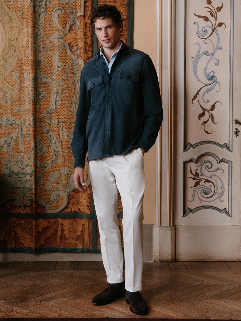 A man wears a dark blue zip cardigan over a white shirt with white trousers.