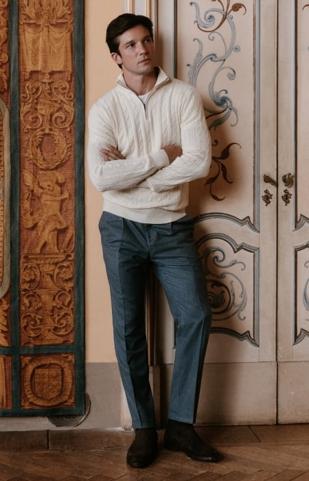 A man wears a white cable-knit quarter-zip sweater with charcoal tailored trousers.
