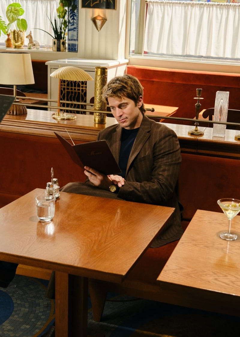 Lucas Bravo seated at a banquette table reading a menu in a brown pinstripe jacket, with a martini glass on the table beside him