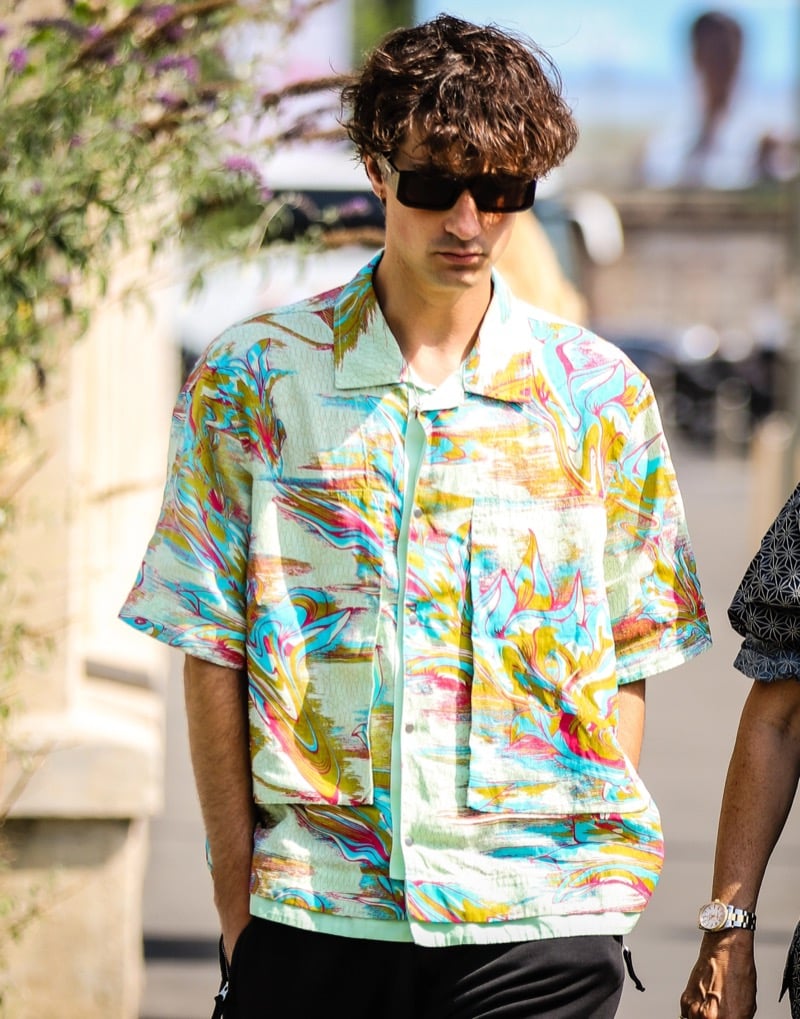 Man in a short-sleeve shirt with a bold multicolor print, black sunglasses and black shorts