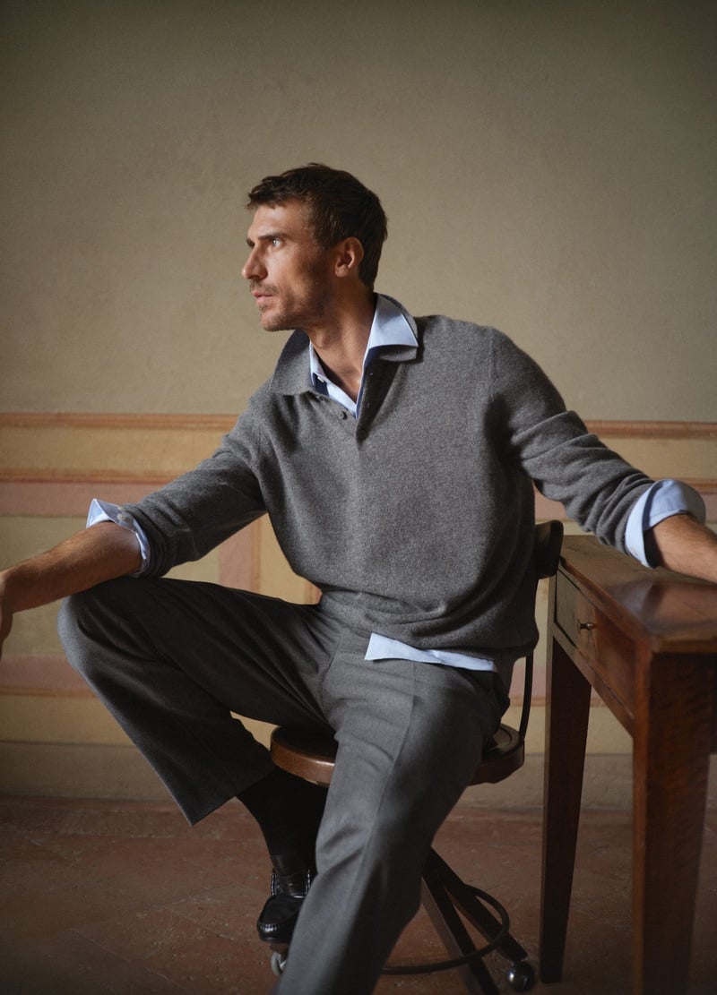 Clément Chabernaud wears a gray knit polo over a blue shirt with tailored trousers while seated in a wooden chair.