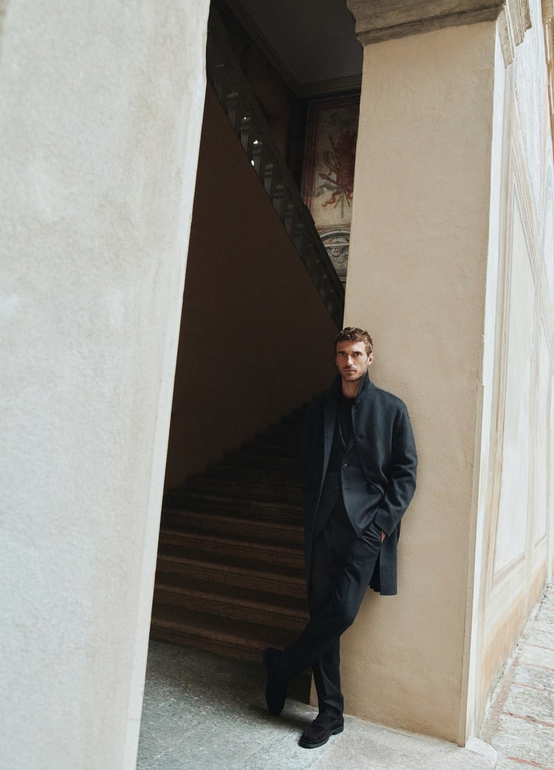Clément Chabernaud stands beside a stone staircase in a dark coat and tailored separates.