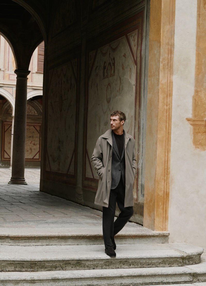 Clément Chabernaud wears a trench coat over tailoring beneath a stone arcade.