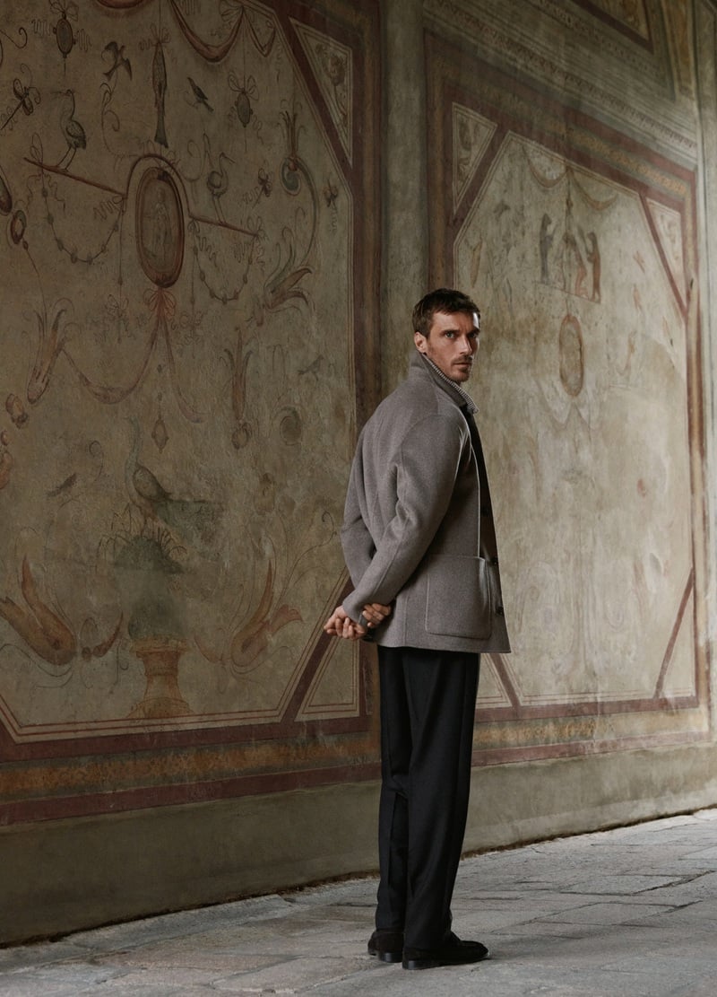 Clément Chabernaud looks over his shoulder in a taupe coat layered over a ribbed zip cardigan.