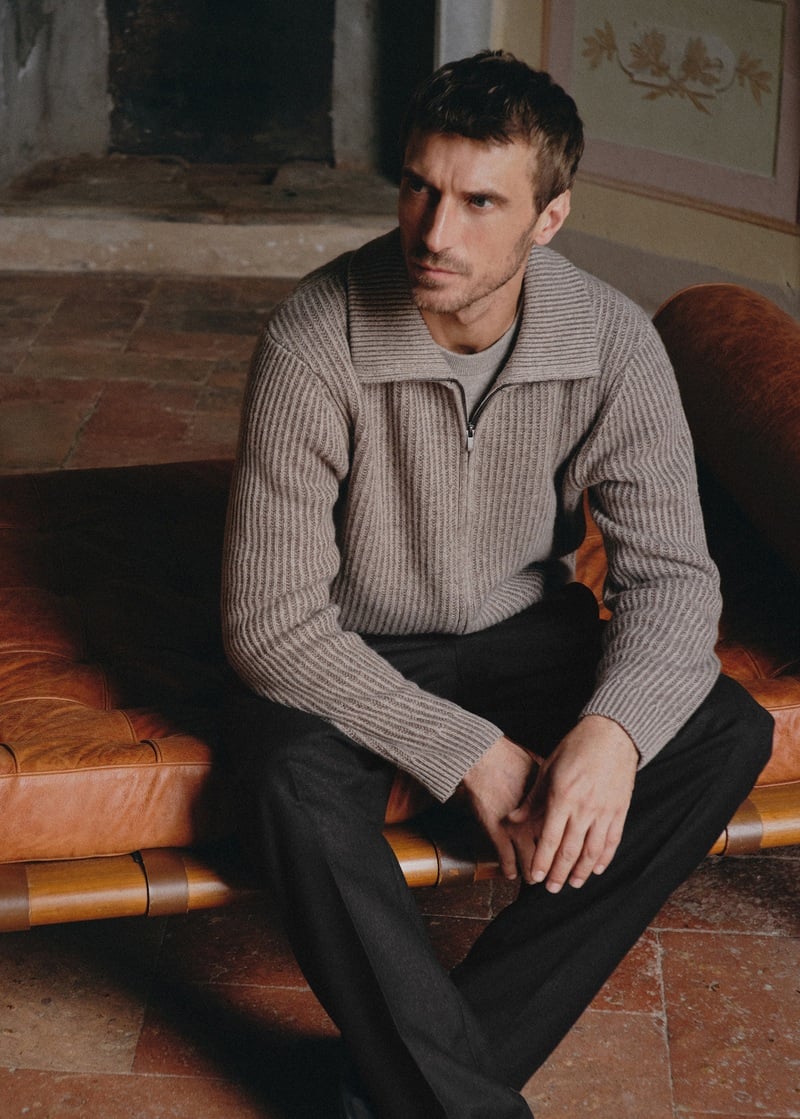 Clément Chabernaud sits on a leather bench in a ribbed zip cardigan and dark trousers.