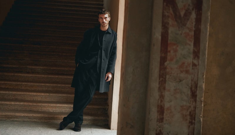 Clément Chabernaud leans against a doorway in a dark coat beside a stone staircase.