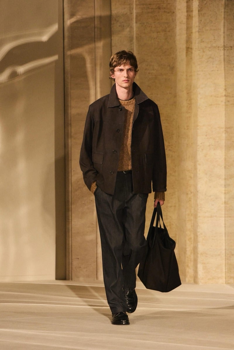 Massimo Dutti Limited Edition fall-winter 2026: Model walks in a dark brown jacket, ochre sweater, and wide charcoal trousers, carrying a black tote.
