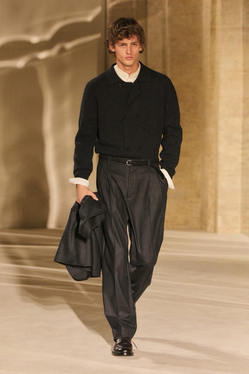 Massimo Dutti Limited Edition fall-winter 2026: Model wears a dark cardigan over a white shirt with charcoal pleated trousers, carrying a jacket.