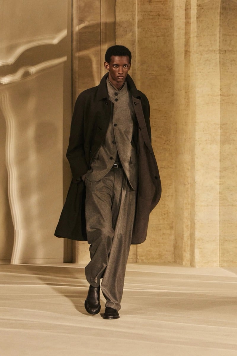 Massimo Dutti Limited Edition fall-winter 2026: Model walks in a dark overcoat over a taupe buttoned knit with matching pleated trousers.
