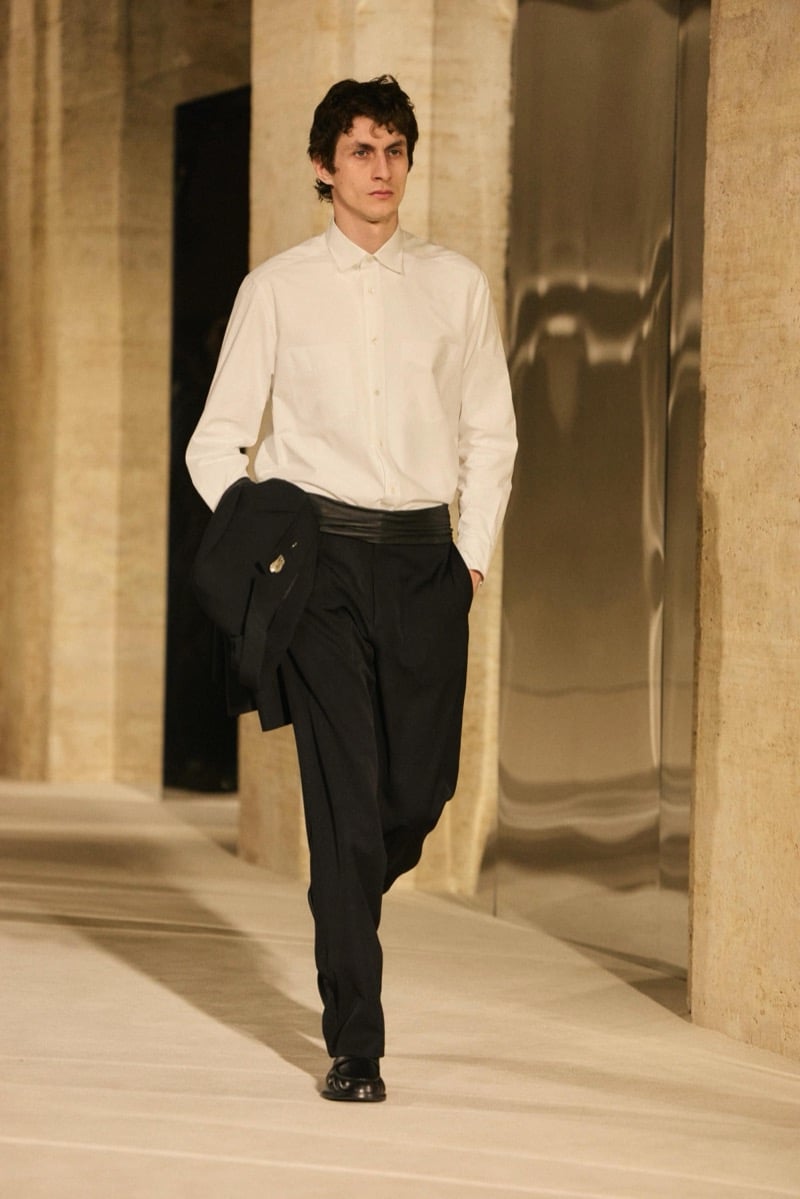 Massimo Dutti Limited Edition fall-winter 2026: Model walks in a white shirt and black pleated trousers with a jacket draped over one arm.